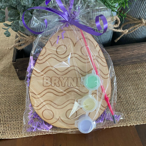 Wooden egg shaped product with personalization wrapped in clear plastic with a purple ribbon on a wooden surface.