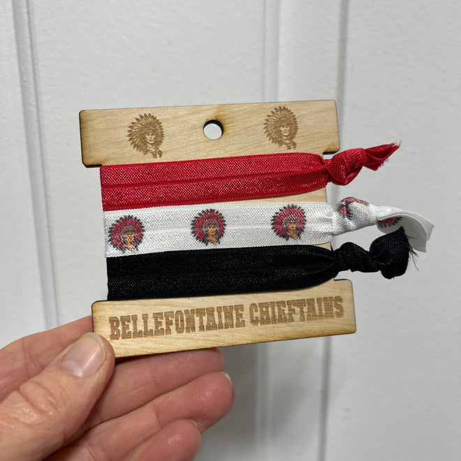 Three hair ties in red, white, and black hanging on a wooden block with branding, held by a hand against a white background.