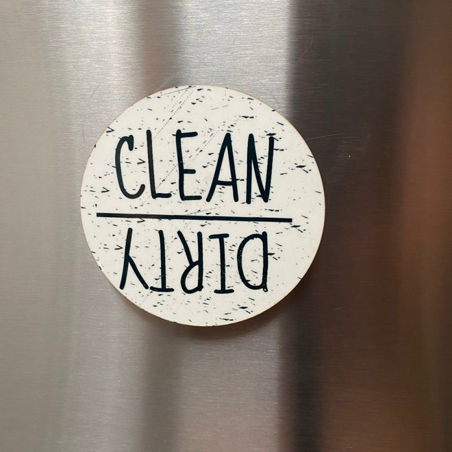 A round white magnetic sticker with black speckled texture and the words 'CLEAN' and 'DIRTY' upside down, shown on a brushed stainless steel surface.