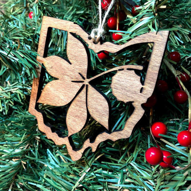 A wooden Christmas ornament shaped like the state of Ohio with a cherry blossom leaf cutout, hanging from a string on a green pine tree with red berries.